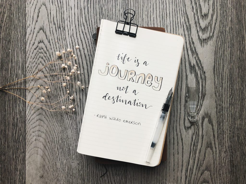 In the centre is a lined notebook page with a hand lettered quote: "Life is a journey not a destination" Underneath the quote is lettered Ralph Waldo Emerson. On the right edge of the page is a clear fountain pen. On the left of the page is a sprig of baby's breath. the backdrop is grey weathered floorboards.