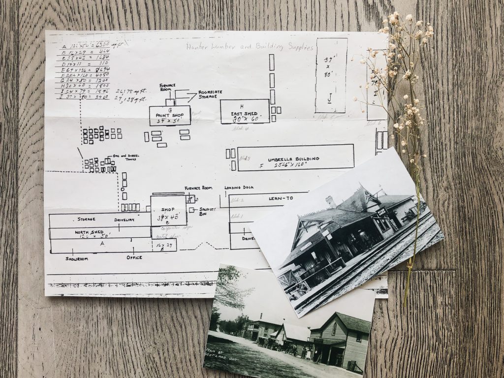 A black and white hand-drawn map of the lumberyard buildings is in the center. Two black and white photos of the Scotland train station and a street view of the village are in the bottom right.  A prig of baby's breath is at the top right. The backdrop is grey farmhouse style floorboards.