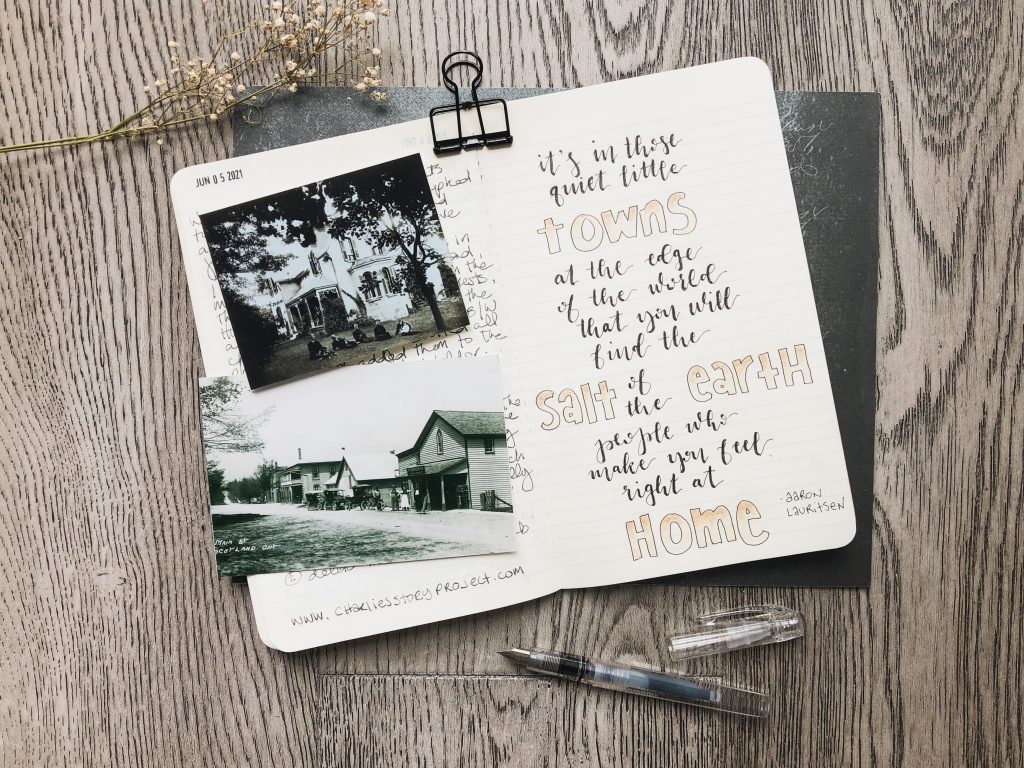 A moleskine notebook is open in the center.  On the left page are two vintage black and white photos depicting a house and a village street. A quote is hand lettered on the right page: "It's in those quiet little towns at the edge of the world that you will find the salt of the earth people who make you feel right at home". The author's name Aaron Lauritsen is in small lettering at the bottom right of the page. An open fountain pen is at the bottom. A sprig of baby's breath is at the top left. The backdrop is grey farmhouse style floorboards.