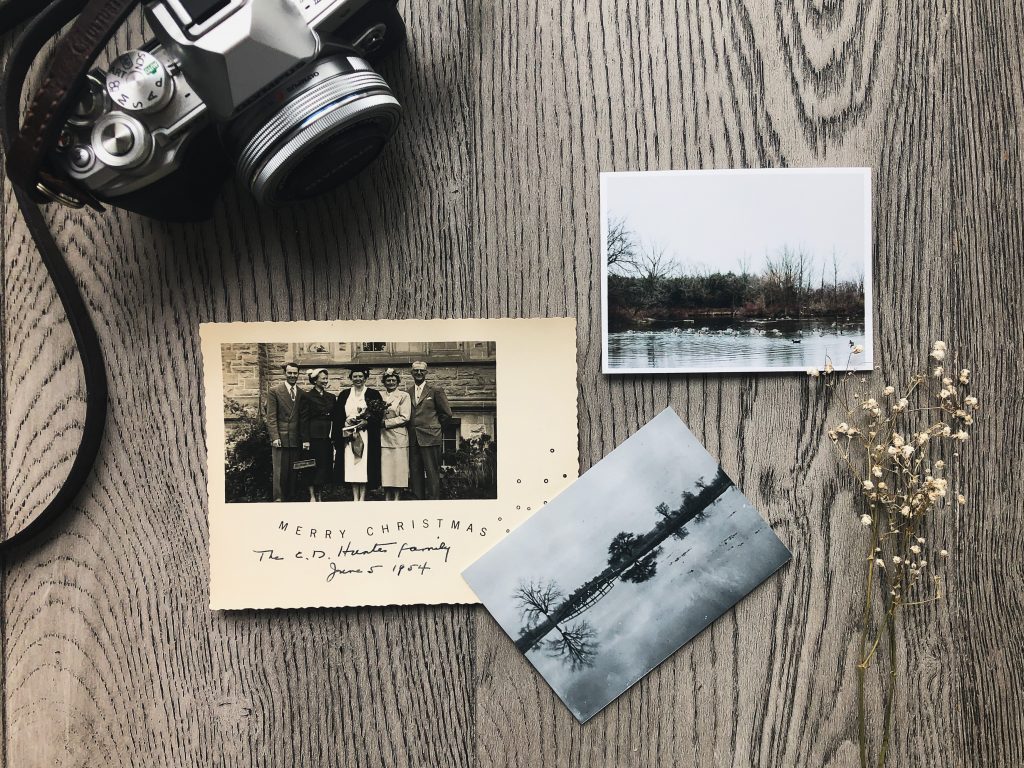 Three photographs are in the centre. On the left is a black and white family photo/Christmas card from 1954. On the right are two photos with scenic views of the pond. A sprig of baby's breath is in the bottom right corner. An Olympus OMD camera is in the top left corner. The backdrop is grey farmhouse style floorboards.