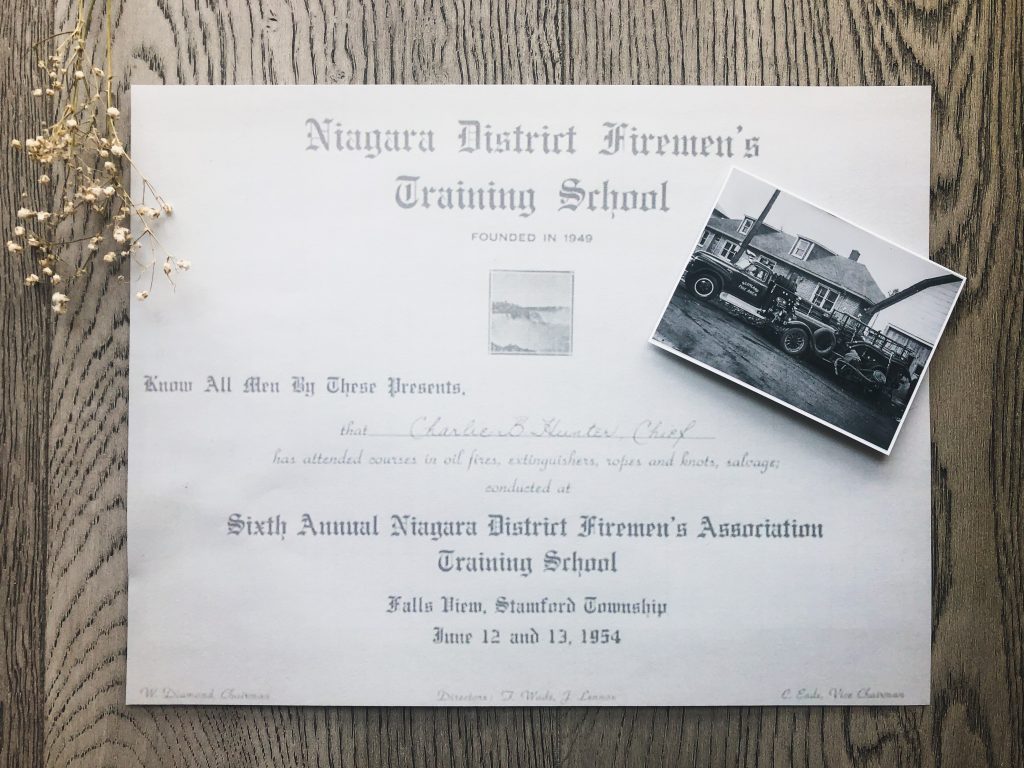 In the centre is a certificate with the title "Niagara District Firemen's Training School" made out to Charlie B. Hunter, Chief.  On the right side of the certificate is a black and white photo of a vintage fire truck. A sprig of baby's breath is at the top left. The backdrop is grey farmhouse style floorboards.