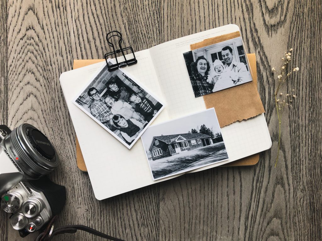A blank notebook is open in the center.  Three black and white photographs are set on top of the blank pages. Two early family snapshots and one of the house. An Olympus OMD camera is in the bottom left corner. The backdrop is grey farmhouse style floorboards.