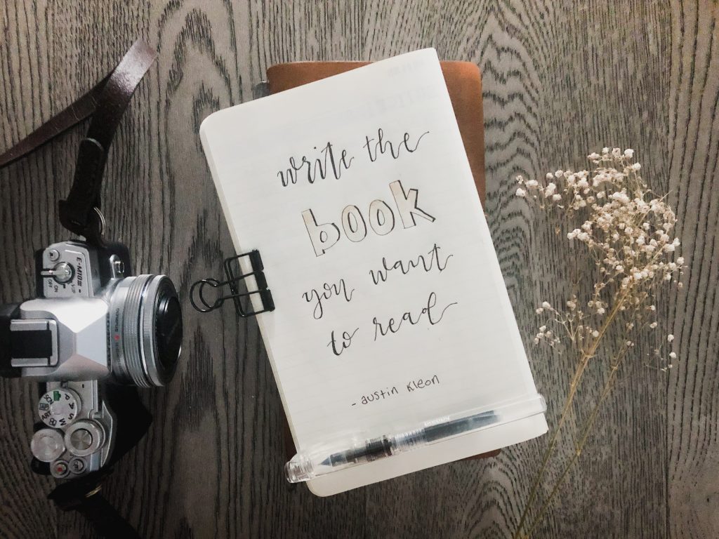 Hand lettering on a notebook page reads "Write the book you want to read". In small writing "Austin Kleon" is underneath the quote. to the left of the notebook is an Olympus OM-D camera. To the right  is a sprig of baby's breath. The backdrop is grey farmhouse style floor boards.